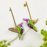 A pair of colorful, handcrafted Hummingbird Earrings with geometric patterns and gold wings, displayed on a white fabric background with green foliage in the foreground.