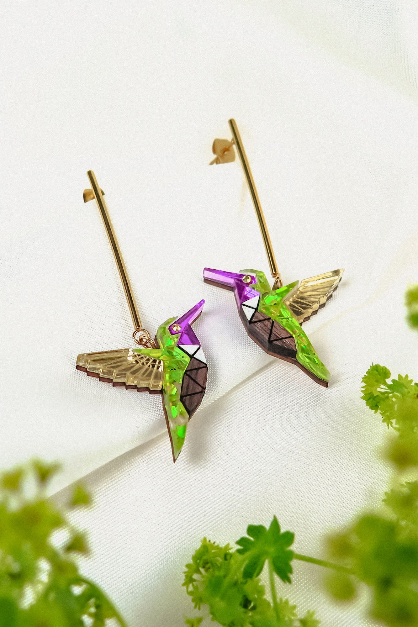 A pair of colorful, handcrafted Hummingbird Earrings with geometric patterns and gold wings, displayed on a white fabric background with green foliage in the foreground.