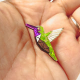 A hand holds a small, colorful Hummingbird Pin shaped like a geometric bird, featuring bright green, purple, and metallic segments with white and brown details—a nature-inspired design celebrating vibrant style.