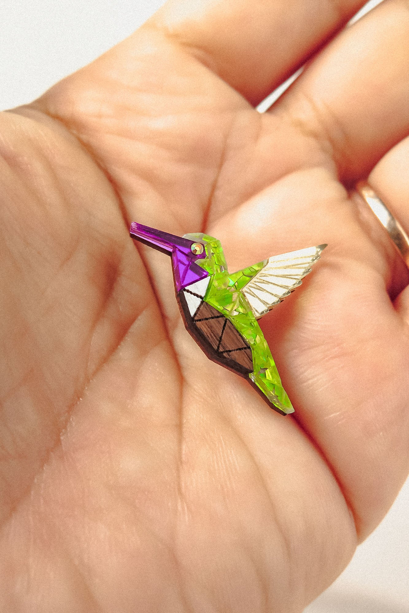 A hand holds a small, colorful Hummingbird Pin shaped like a geometric bird, featuring bright green, purple, and metallic segments with white and brown details—a nature-inspired design celebrating vibrant style.
