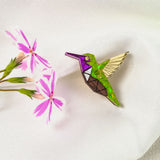 A colorful Hummingbird Pin with green, purple, brown, and gold sections rests on white fabric next to a sprig of pink flowers, showcasing nature-inspired design and timeless charm.