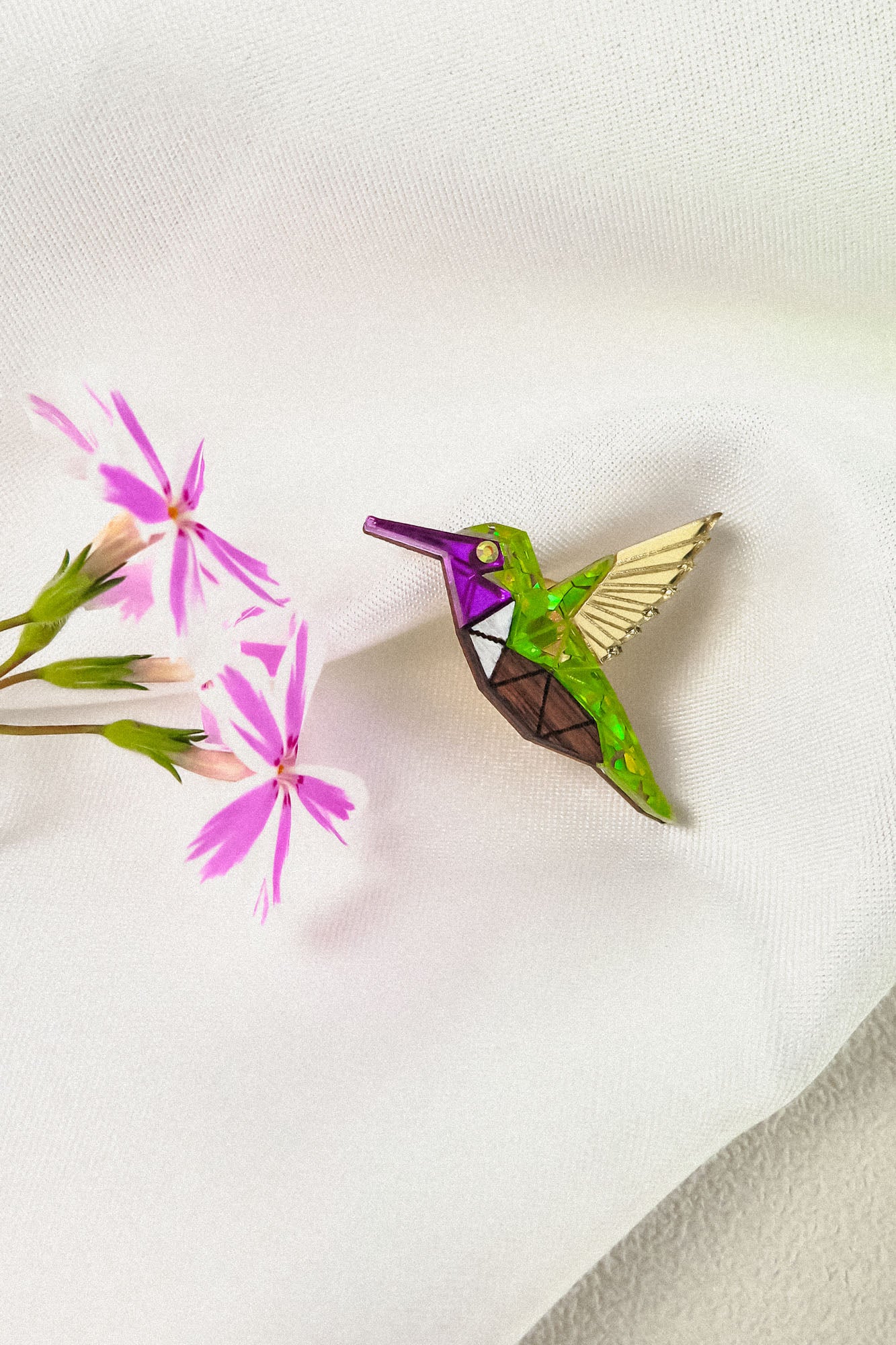 A colorful Hummingbird Pin with green, purple, brown, and gold sections rests on white fabric next to a sprig of pink flowers, showcasing nature-inspired design and timeless charm.