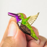 A hand holds a small, colorful hummingbird pin featuring a geometric, nature-inspired design with green, purple, gold, and brown sections and transparent wings.