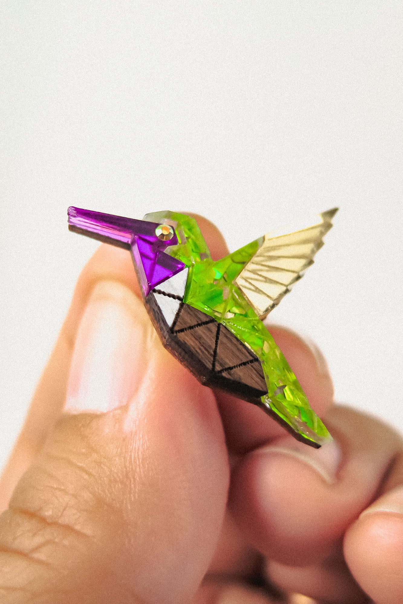 A hand holds a small, colorful hummingbird pin featuring a geometric, nature-inspired design with green, purple, gold, and brown sections and transparent wings.
