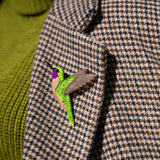 A close-up of a handcrafted hummingbird brooch, made from premium materials, pinned to a houndstooth-patterned coat with a green turtleneck sweater visible underneath.