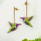 Two colorful Hummingbird Earrings with geometric designs and long gold posts rest on a white fabric background, showcasing handcrafted details, with green foliage blurred in the foreground.