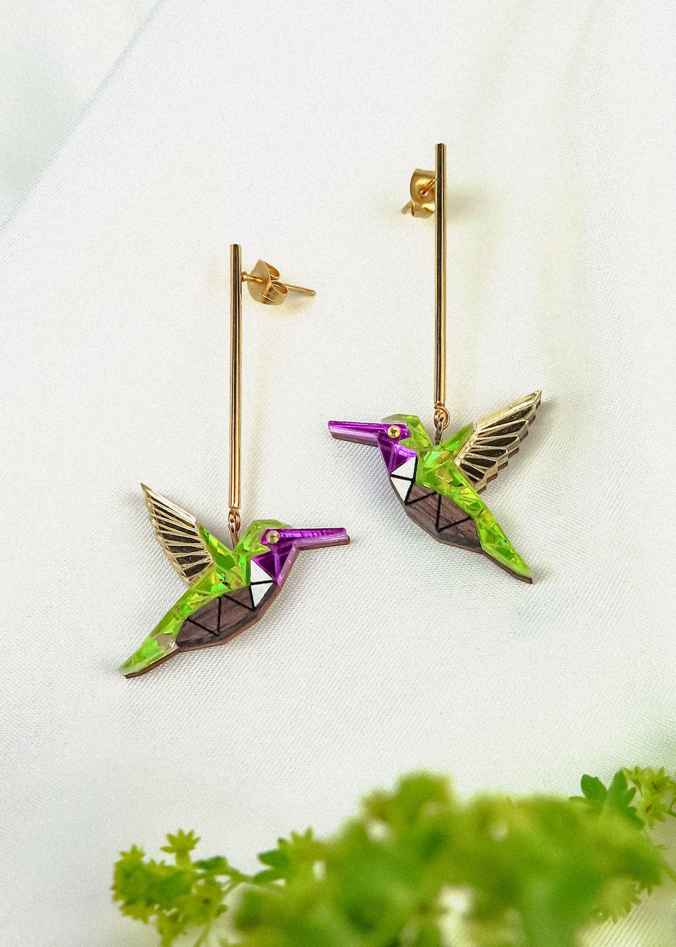 Two colorful Hummingbird Earrings with geometric designs and long gold posts rest on a white fabric background, showcasing handcrafted details, with green foliage blurred in the foreground.