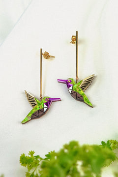 Two colorful Hummingbird Earrings with geometric designs and long gold posts rest on a white fabric background, showcasing handcrafted details, with green foliage blurred in the foreground.