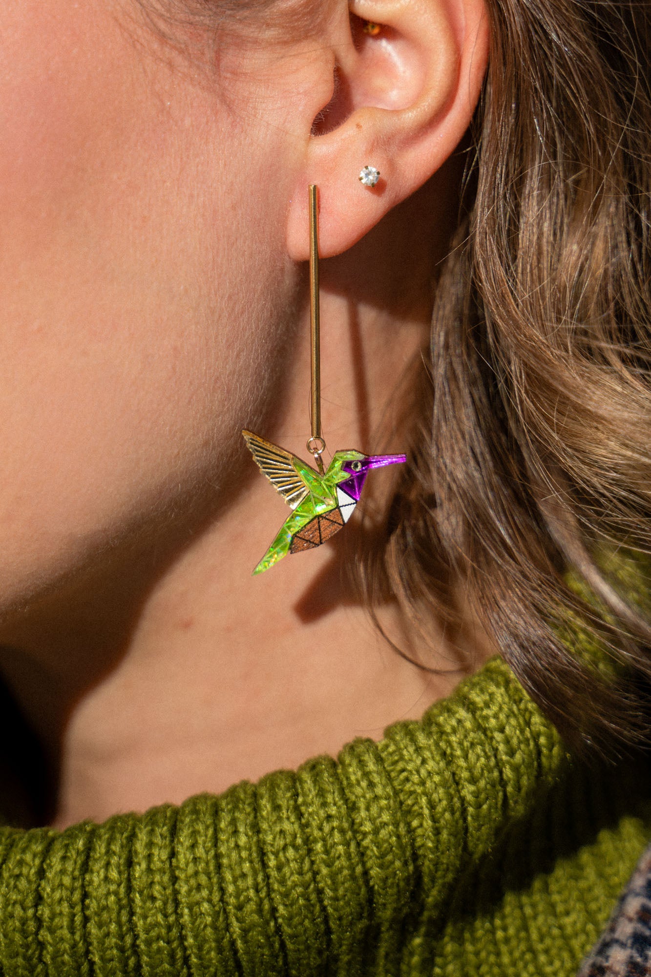 Close-up of a person’s ear wearing long, colorful handcrafted Hummingbird Earrings and a small stud earring. The person has light brown hair and is wearing a green knit sweater, showcasing sustainable craftsmanship.