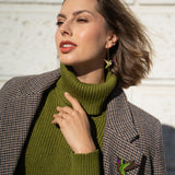 Woman wearing a green turtleneck sweater and plaid coat, accessorized with handcrafted Hummingbird Earrings and a brooch, poses outdoors against a light textured wall, touching her collar and looking off to the side.