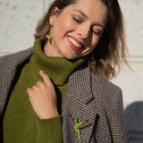 A smiling woman in a green turtleneck and plaid jacket stands against a light wall, wearing handcrafted Hummingbird Earrings and matching lapel jewelry. Sunlight highlights her face as she closes her eyes, radiating joy and sustainable craftsmanship.