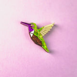 A colorful, geometric Hummingbird Pin featuring purple, green, brown, and yellow sections lies on a pastel pink background—showcasing nature-inspired design with vibrant elegance.