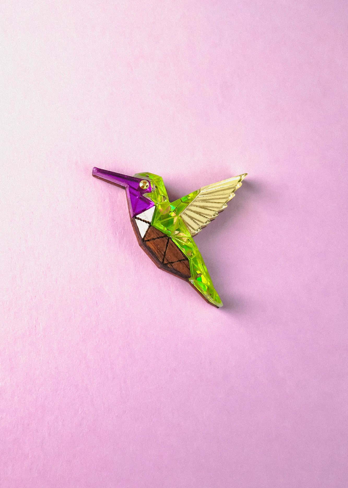 A colorful, geometric Hummingbird Pin featuring purple, green, brown, and yellow sections lies on a pastel pink background—showcasing nature-inspired design with vibrant elegance.