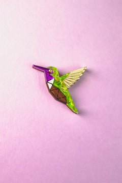 A colorful, geometric Hummingbird Pin featuring purple, green, brown, and yellow sections lies on a pastel pink background—showcasing nature-inspired design with vibrant elegance.