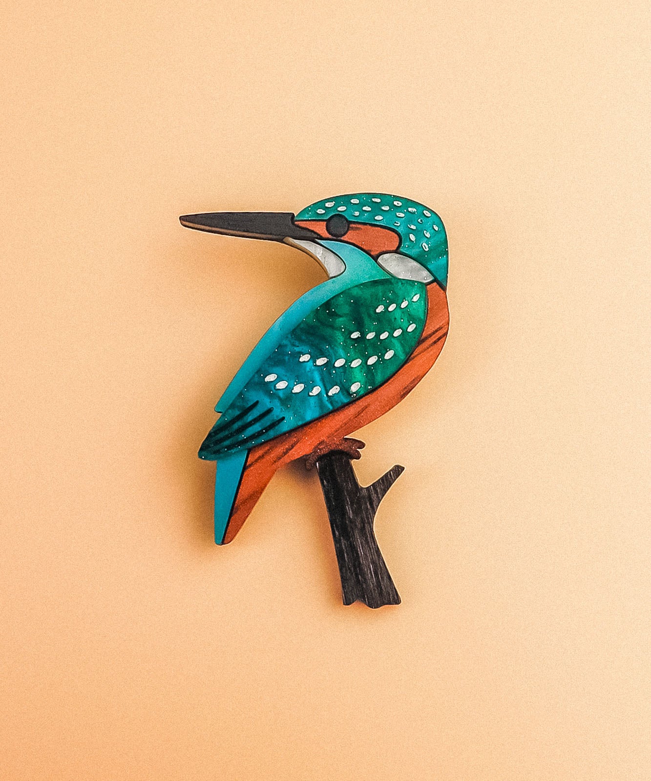 A colorful, stylized Kingfisher Brooch handcrafted from sustainable materials, with teal, orange, and white markings perched on a small black branch, set against a pale peach background.