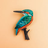 A colorful, stylized Kingfisher Brooch handcrafted from sustainable materials, with teal, orange, and white markings perched on a small black branch, set against a pale peach background.