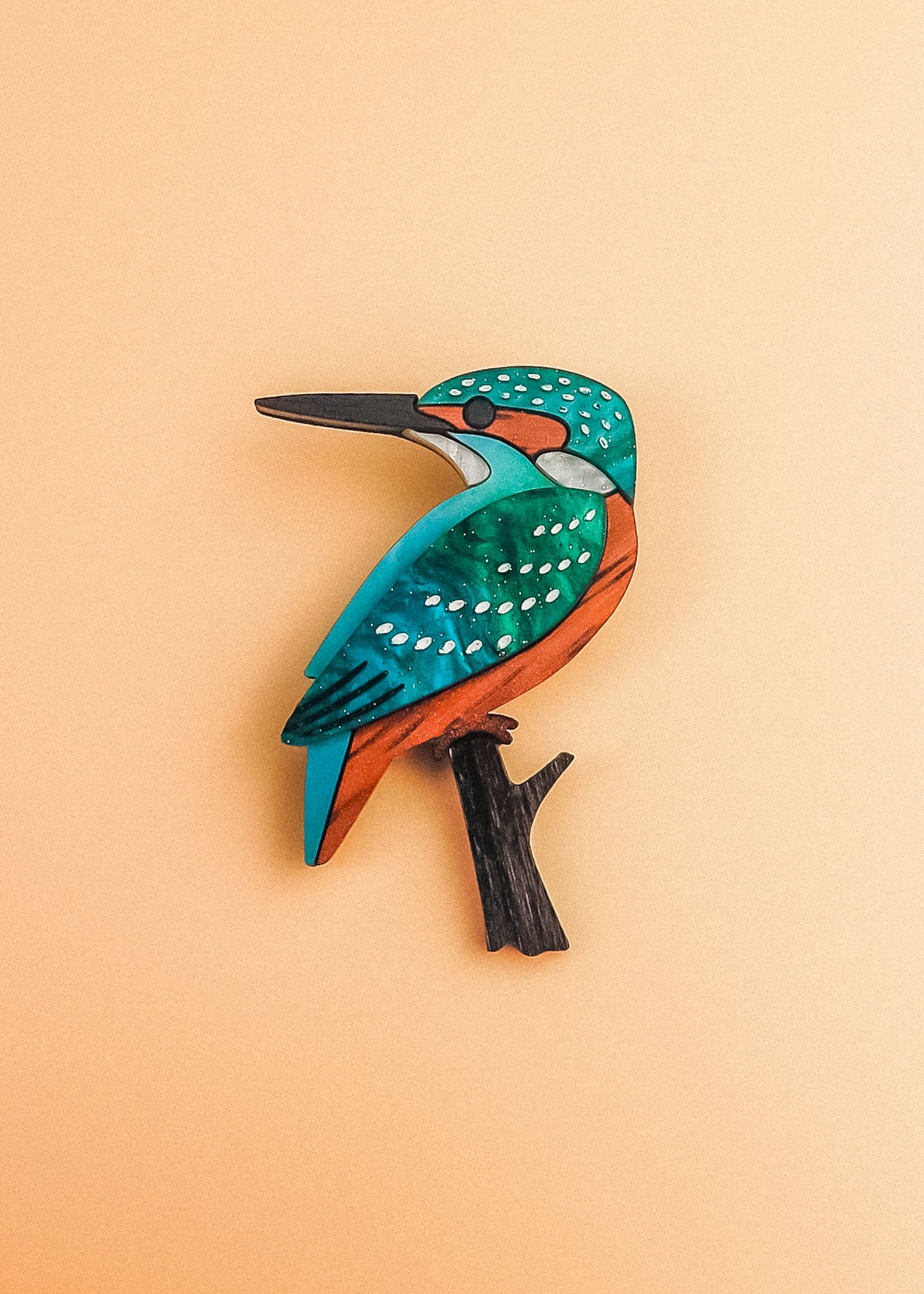 A colorful, stylized Kingfisher Brooch handcrafted from sustainable materials, with teal, orange, and white markings perched on a small black branch, set against a pale peach background.