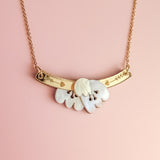 A handcrafted gold chain Tulip Necklace with a curved gold bar and heart engravings, featuring a white, petal-shaped pendant—radiating romantic charm against a light pink background.