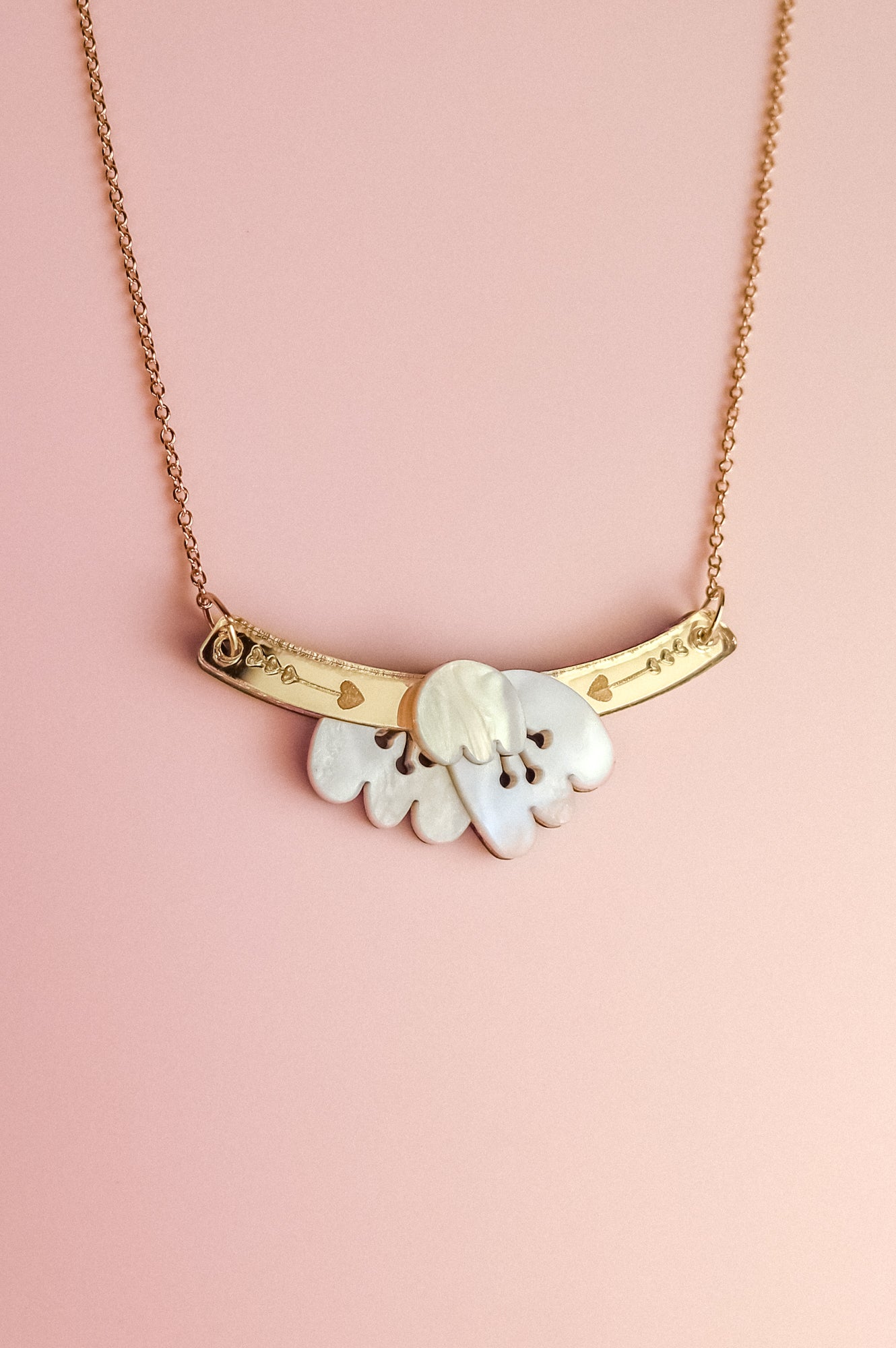 A handcrafted gold chain Tulip Necklace with a curved gold bar and heart engravings, featuring a white, petal-shaped pendant—radiating romantic charm against a light pink background.