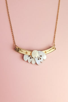A handcrafted gold chain Tulip Necklace with a curved gold bar and heart engravings, featuring a white, petal-shaped pendant—radiating romantic charm against a light pink background.