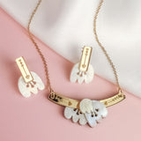 A handcrafted gold Tulip Necklace and matching earrings with white floral-shaped pendants and engraved gold bars are displayed on a soft pink and white fabric background, radiating romantic charm.