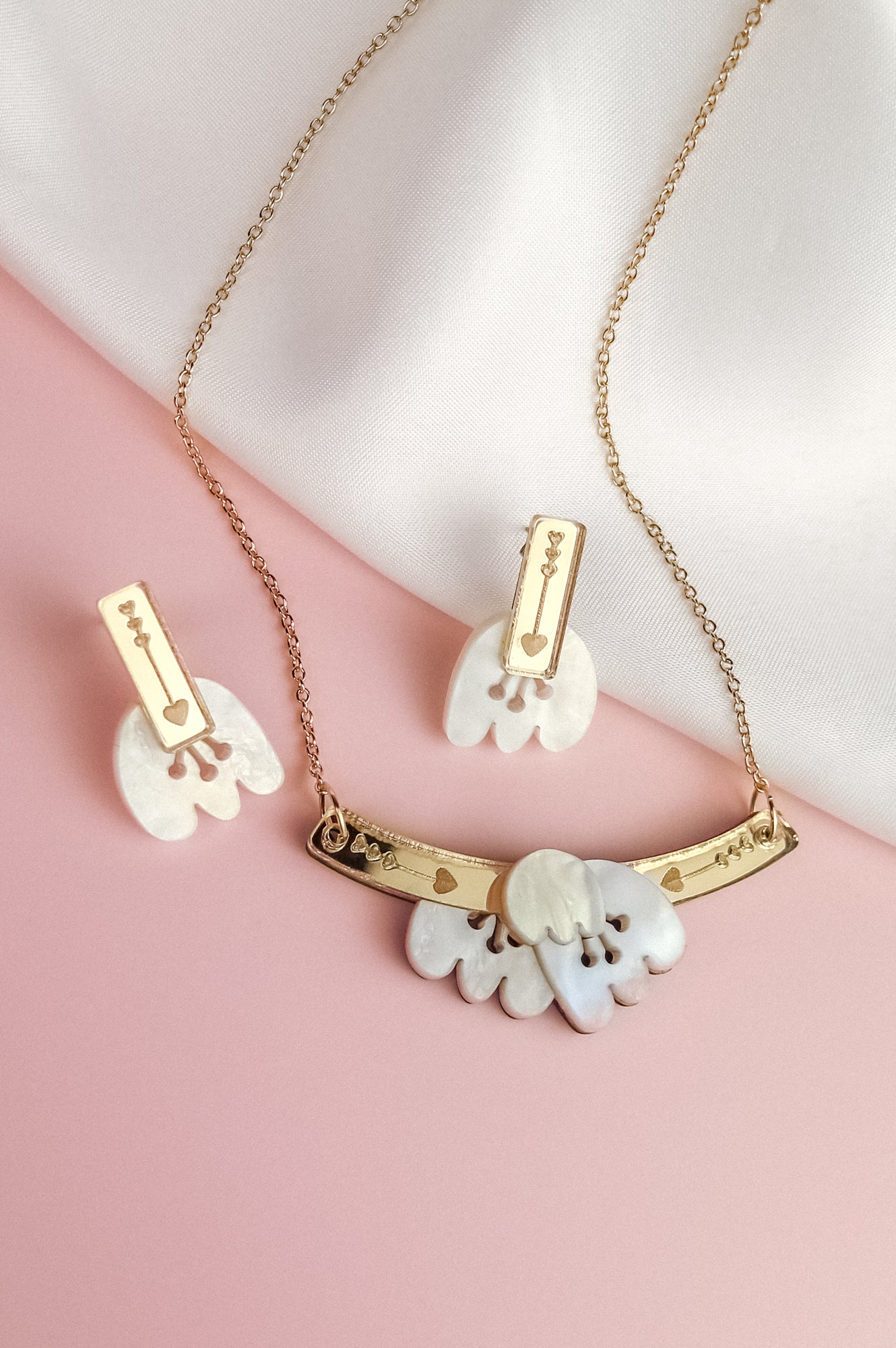 A handcrafted gold Tulip Necklace and matching earrings with white floral-shaped pendants and engraved gold bars are displayed on a soft pink and white fabric background, radiating romantic charm.