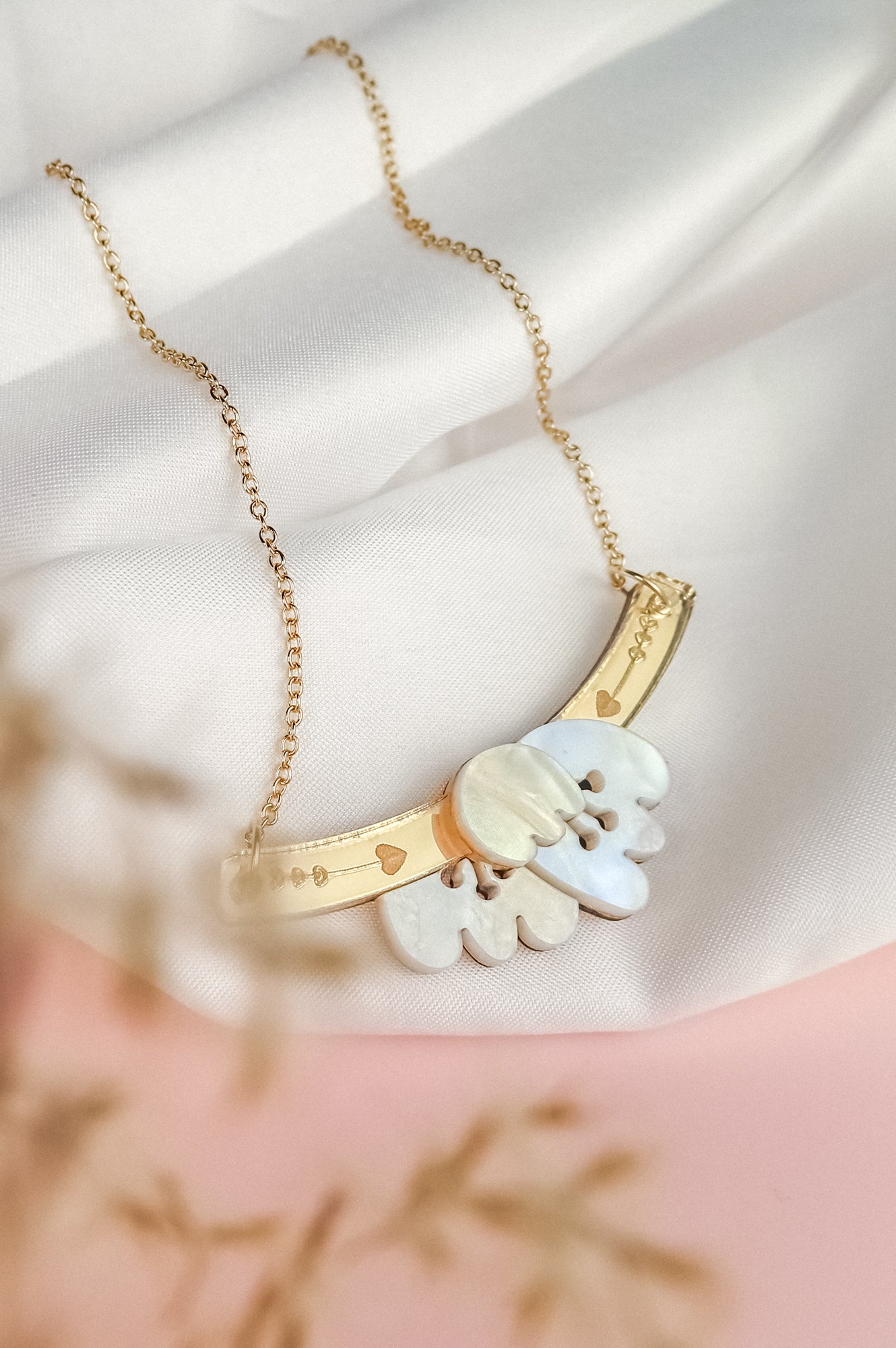 A gold Tulip Necklace with a delicate chain, featuring an intricate pendant with white and gold elements and small heart motifs, exudes romantic charm as it rests on a soft white fabric background.