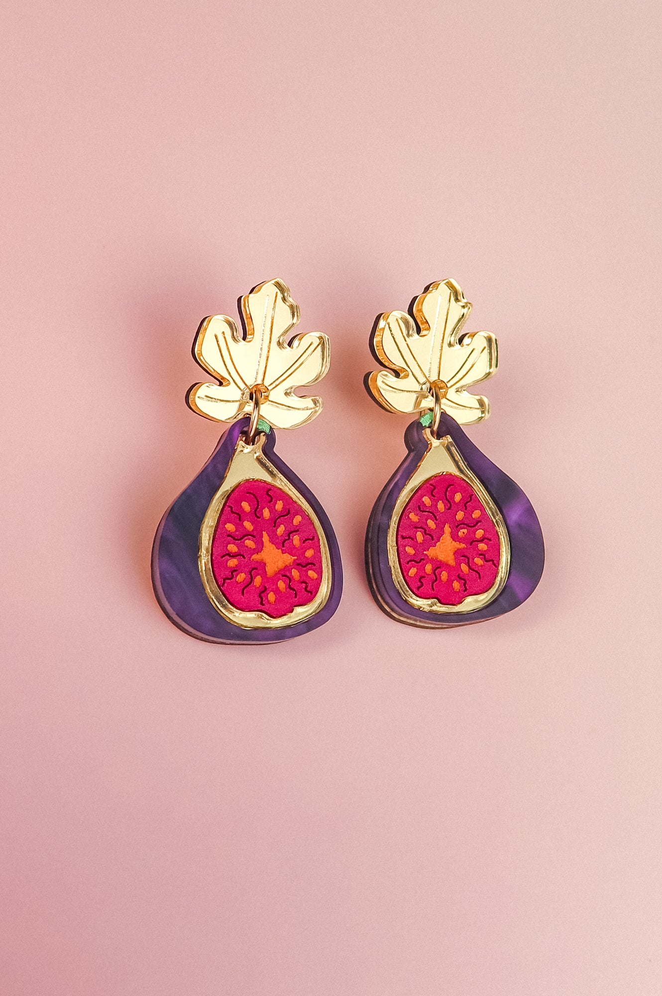Handcrafted Fig Earrings featuring gold mirror leaves, purple outer edges, and bright pink centers, beautifully displayed on a light pink background.