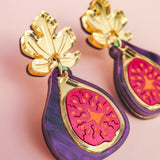 A close-up of handcrafted Fig Earrings shaped like figs, featuring gold mirror leaves at the top, vibrant purple edges, and a bright pink and orange fig center, set against a soft pink background.