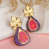 A pair of handcrafted Fig Earrings with gold mirror leaves, purple edges, and a pink center, displayed on a soft white and pink background with dried flowers.