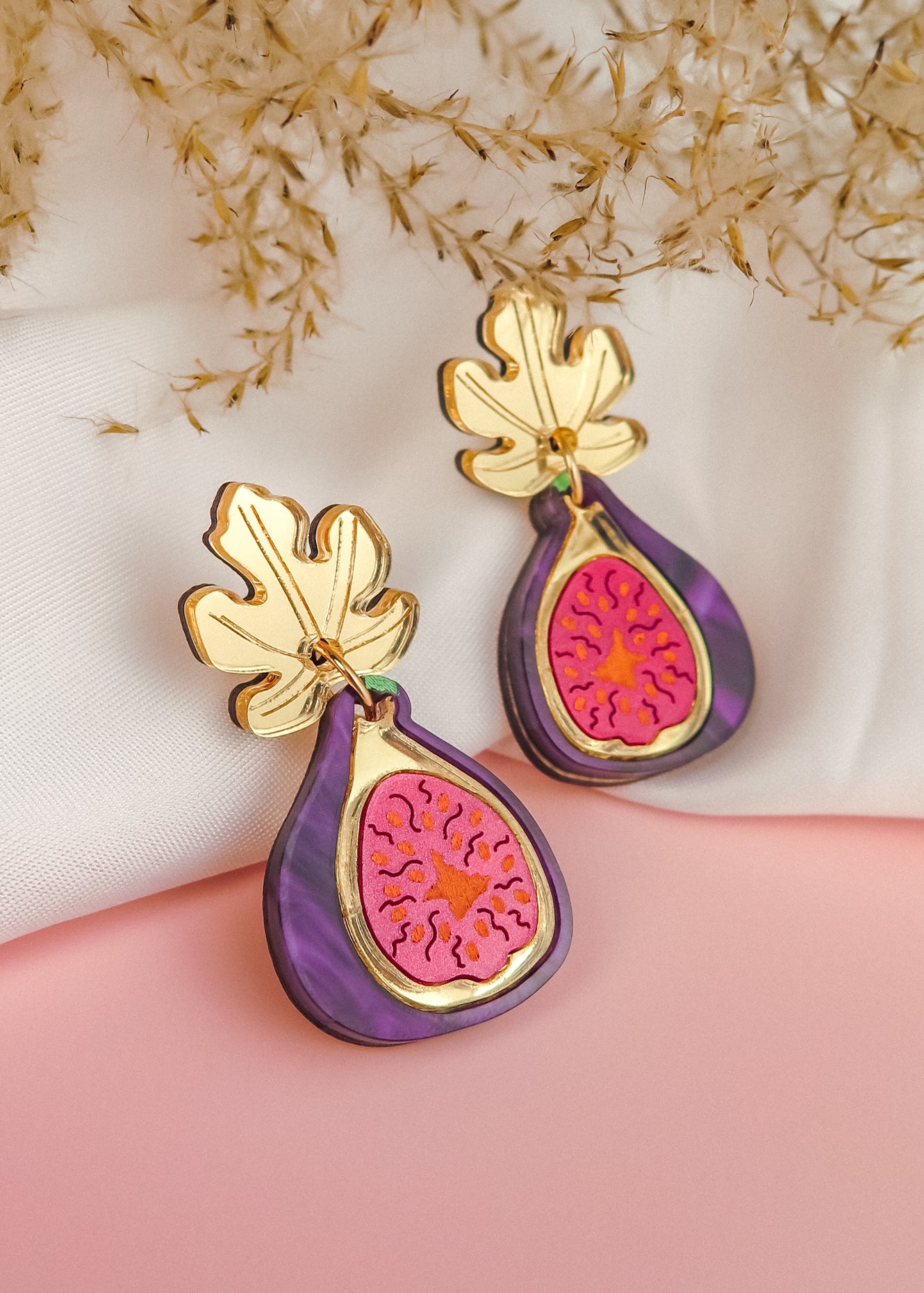A pair of handcrafted Fig Earrings with gold mirror leaves, purple edges, and a pink center, displayed on a soft white and pink background with dried flowers.
