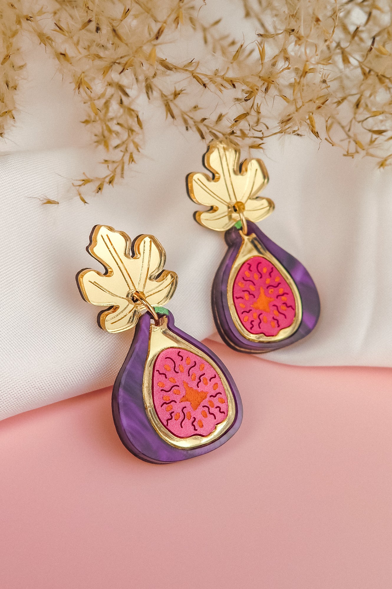 A pair of handcrafted Fig Earrings with gold mirror leaves, purple edges, and a pink center, displayed on a soft white and pink background with dried flowers.