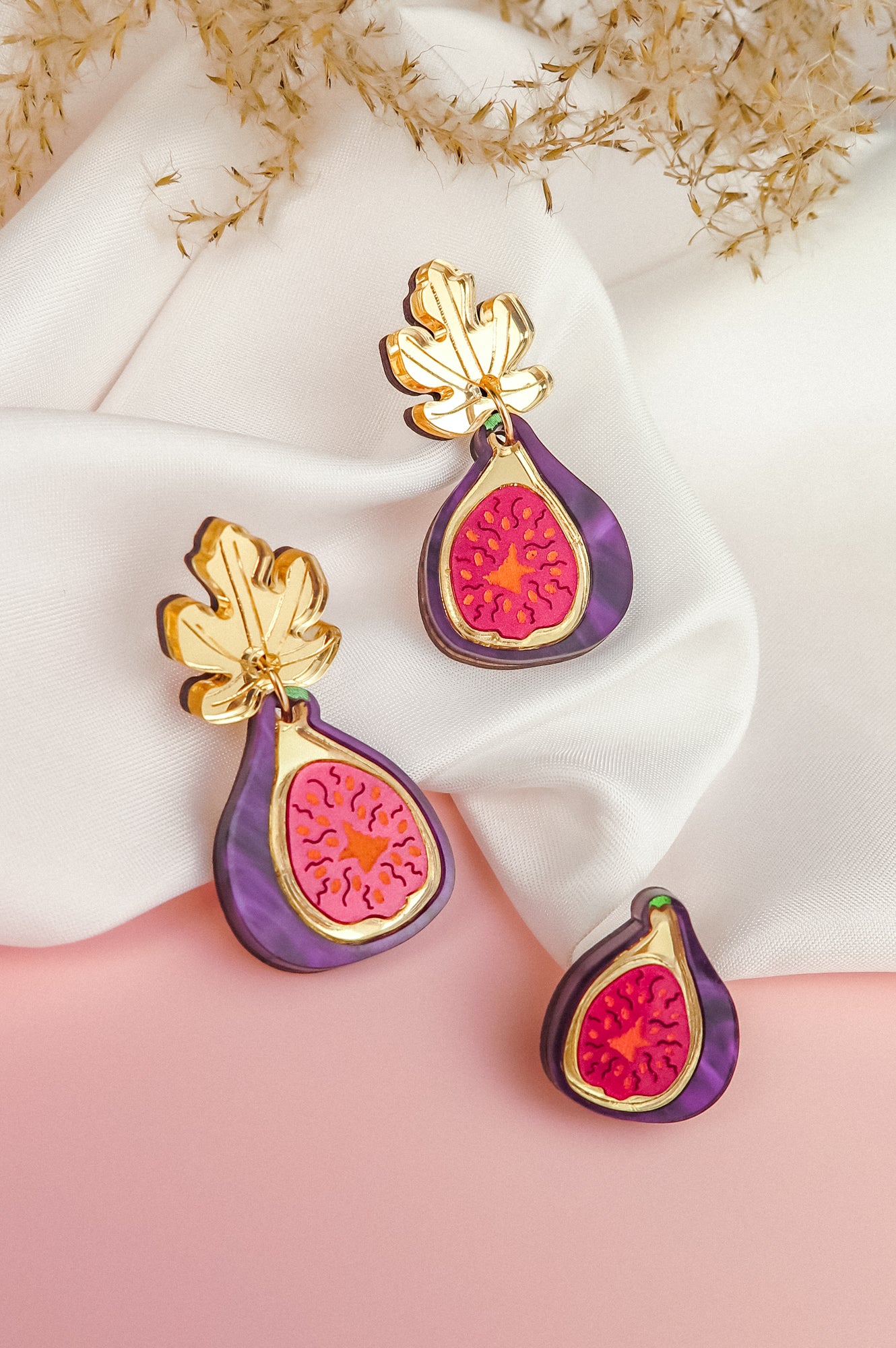 Three handcrafted Fig Earrings, featuring gold mirror leaves and vibrant purple and pink detailing, are displayed on white fabric and a pink surface, with dried foliage in the background.