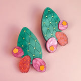 A pair of statement earrings shaped like green opuncia cactus pads with pink and orange flowers, decorated with small yellow dots, displayed on a light pink background.