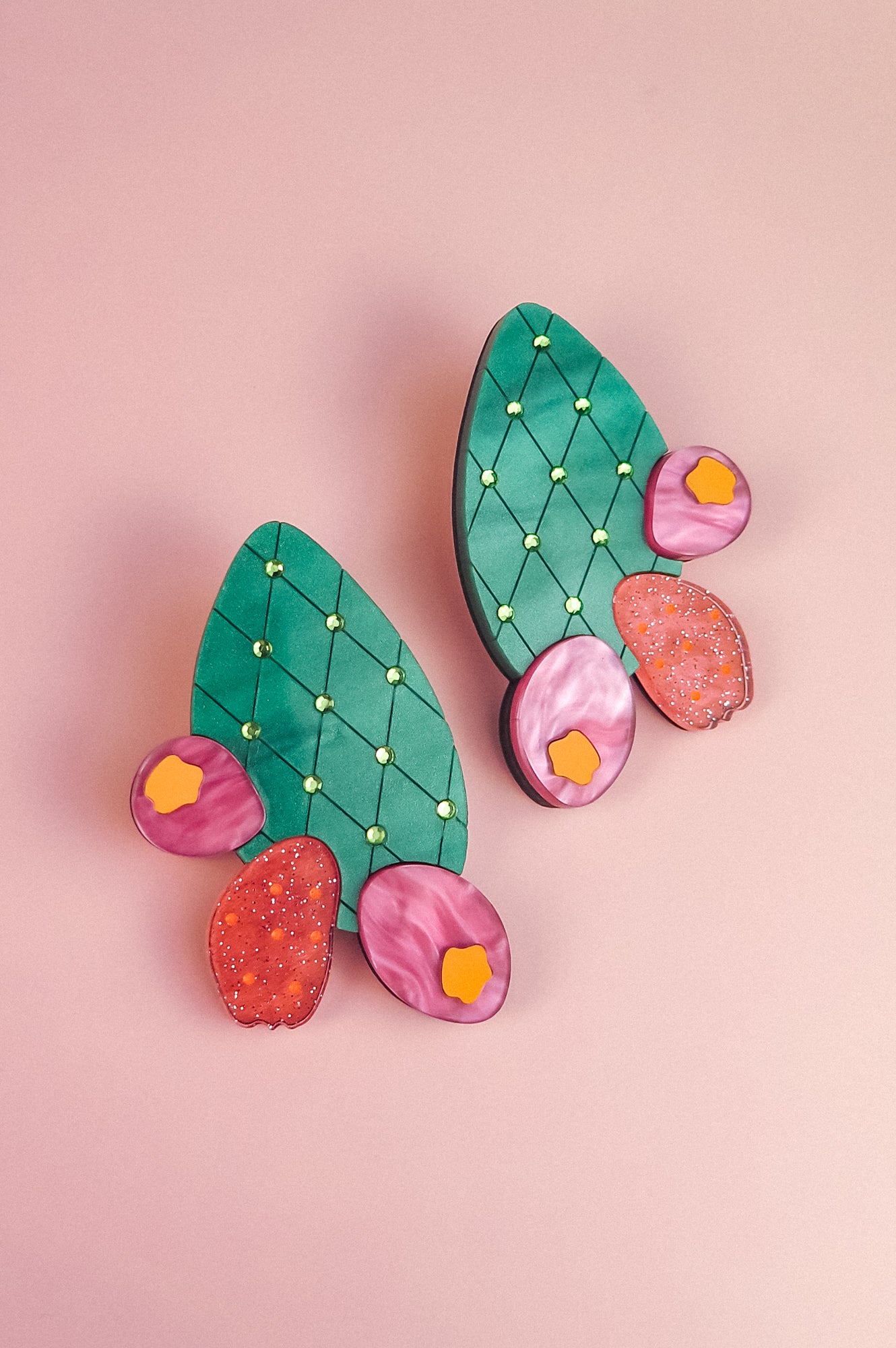 A pair of statement earrings shaped like green opuncia cactus pads with pink and orange flowers, decorated with small yellow dots, displayed on a light pink background.