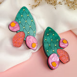 A pair of colorful statement earrings inspired by the opuncia cactus, featuring green segments, yellow dots, and pink and orange flower decorations, displayed on white fabric with dried foliage in the background.
