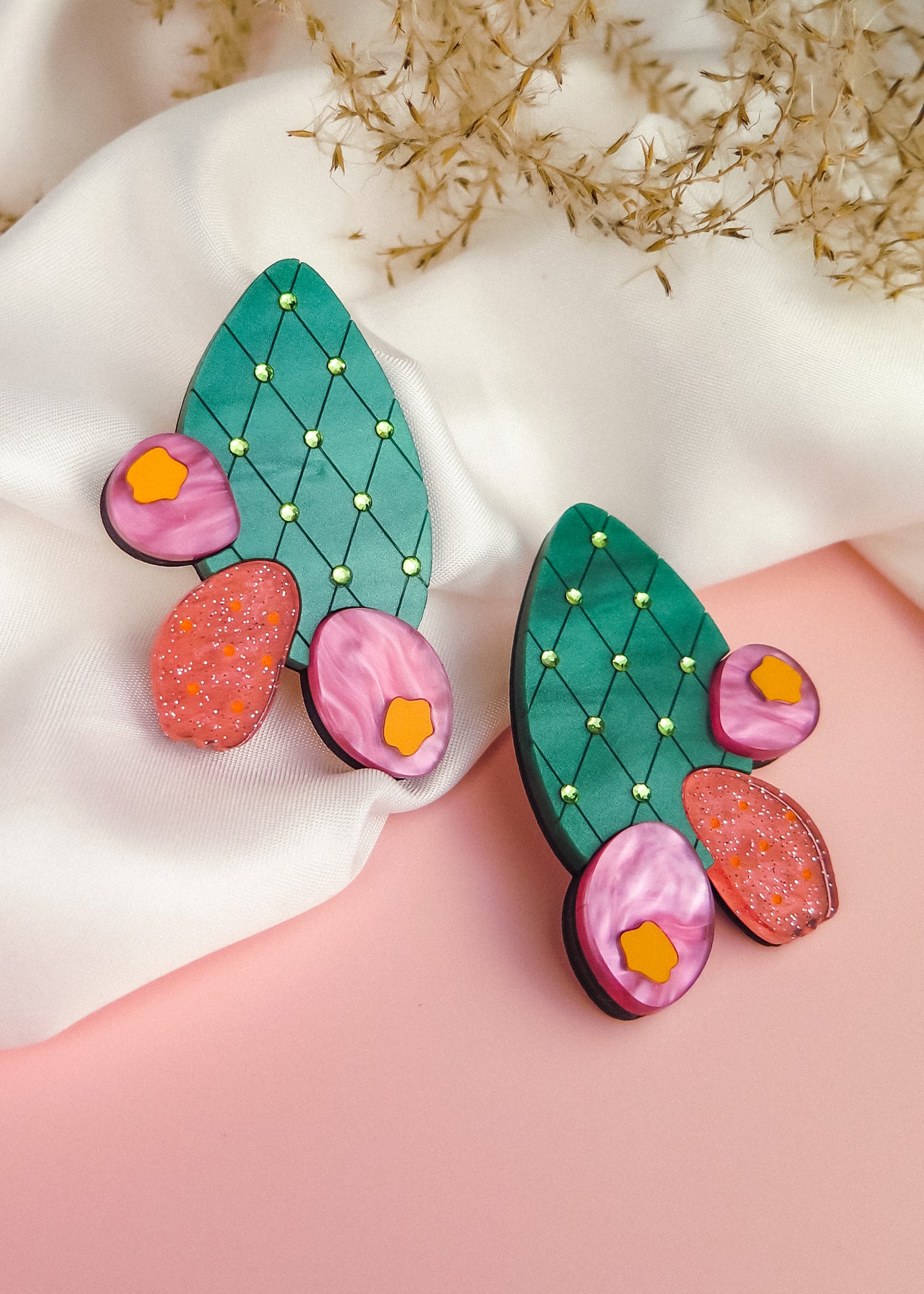 A pair of colorful statement earrings inspired by the opuncia cactus, featuring green segments, yellow dots, and pink and orange flower decorations, displayed on white fabric with dried foliage in the background.