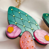 A close-up of bold statement earrings, stylized as green leaves with yellow dots and pink, orange, and purple accents—reminiscent of the opuncia cactus—displayed on soft white fabric with delicate dried flowers above.
