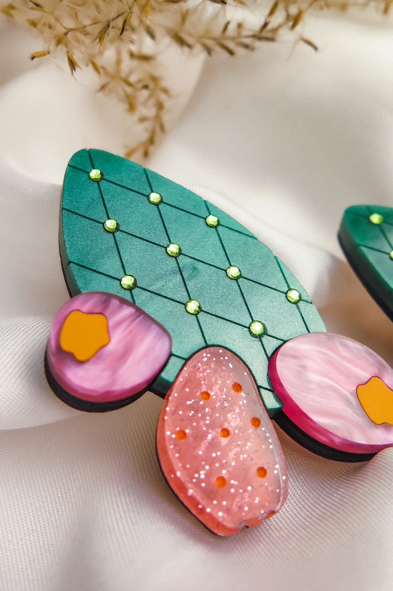 A close-up of bold statement earrings, stylized as green leaves with yellow dots and pink, orange, and purple accents—reminiscent of the opuncia cactus—displayed on soft white fabric with delicate dried flowers above.