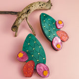 Colorful statement earrings shaped like opuncia cactus pads with green tops, gold dots, and pink and red decorative accents, displayed on a pink background with a piece of dried branch for a bold design touch.