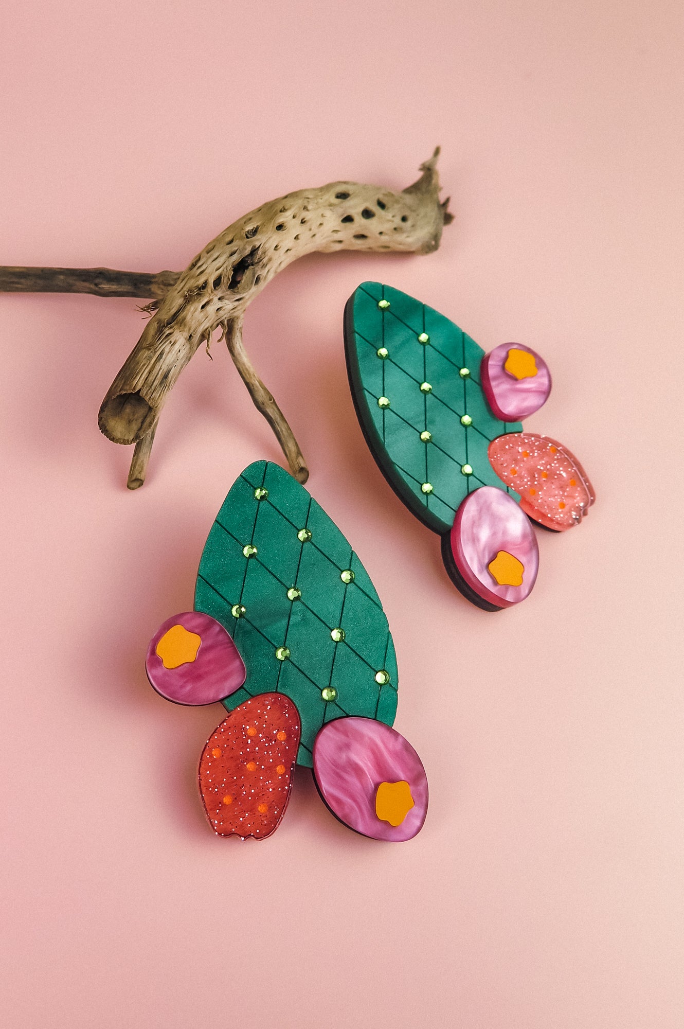 Colorful statement earrings shaped like opuncia cactus pads with green tops, gold dots, and pink and red decorative accents, displayed on a pink background with a piece of dried branch for a bold design touch.