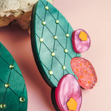 Colorful statement earrings shaped like opuncia cactus pads with a bold design featuring a green diamond pattern, small gems, and round pink and orange accents, displayed on a pink background near a beige rock.