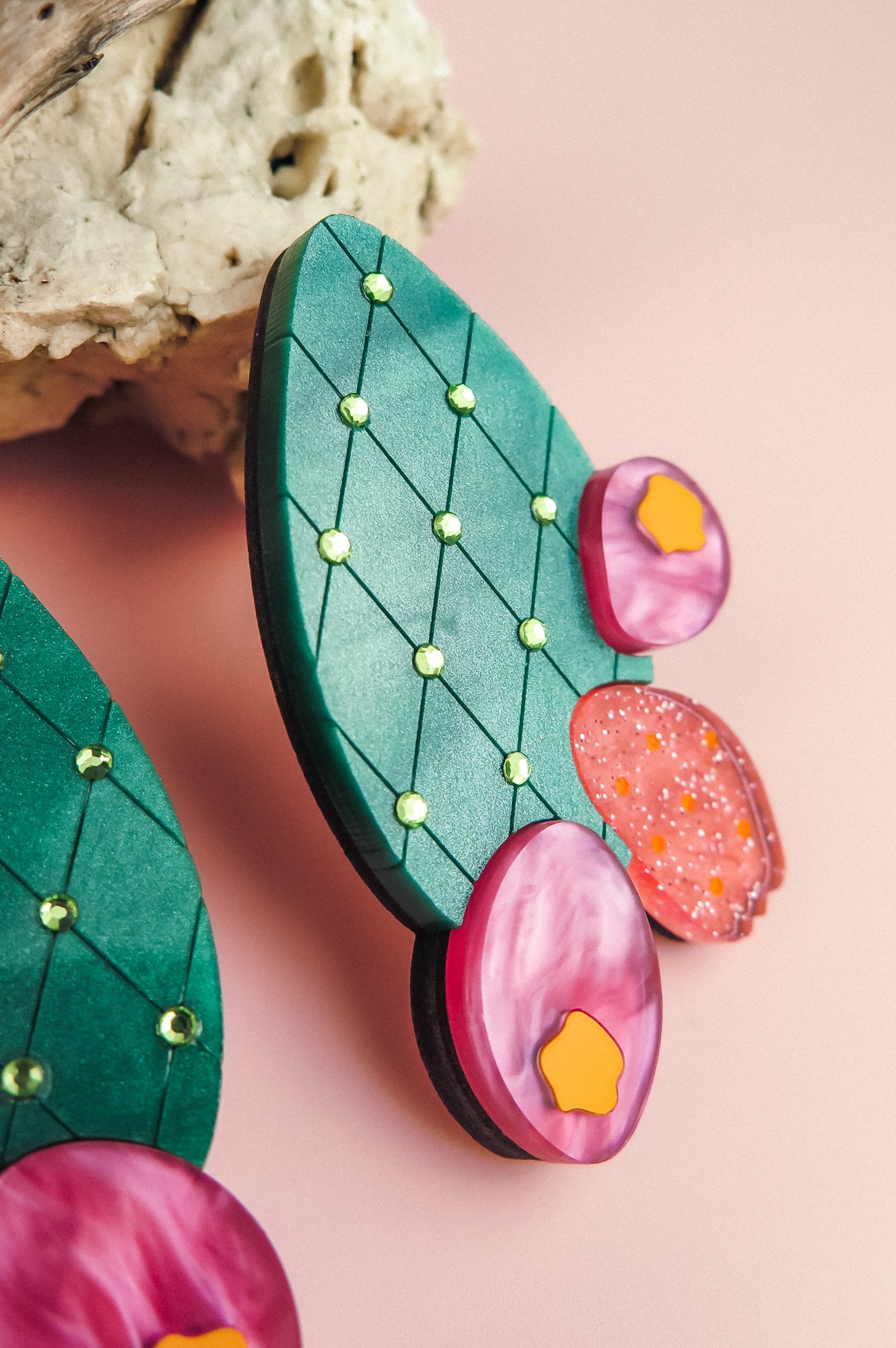 Colorful statement earrings shaped like opuncia cactus pads with a bold design featuring a green diamond pattern, small gems, and round pink and orange accents, displayed on a pink background near a beige rock.