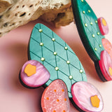 A close-up of colorful, handmade statement earrings shaped like opuncia cactus pads with green, pink, and orange sections, lying on a pink surface next to a textured piece of driftwood.