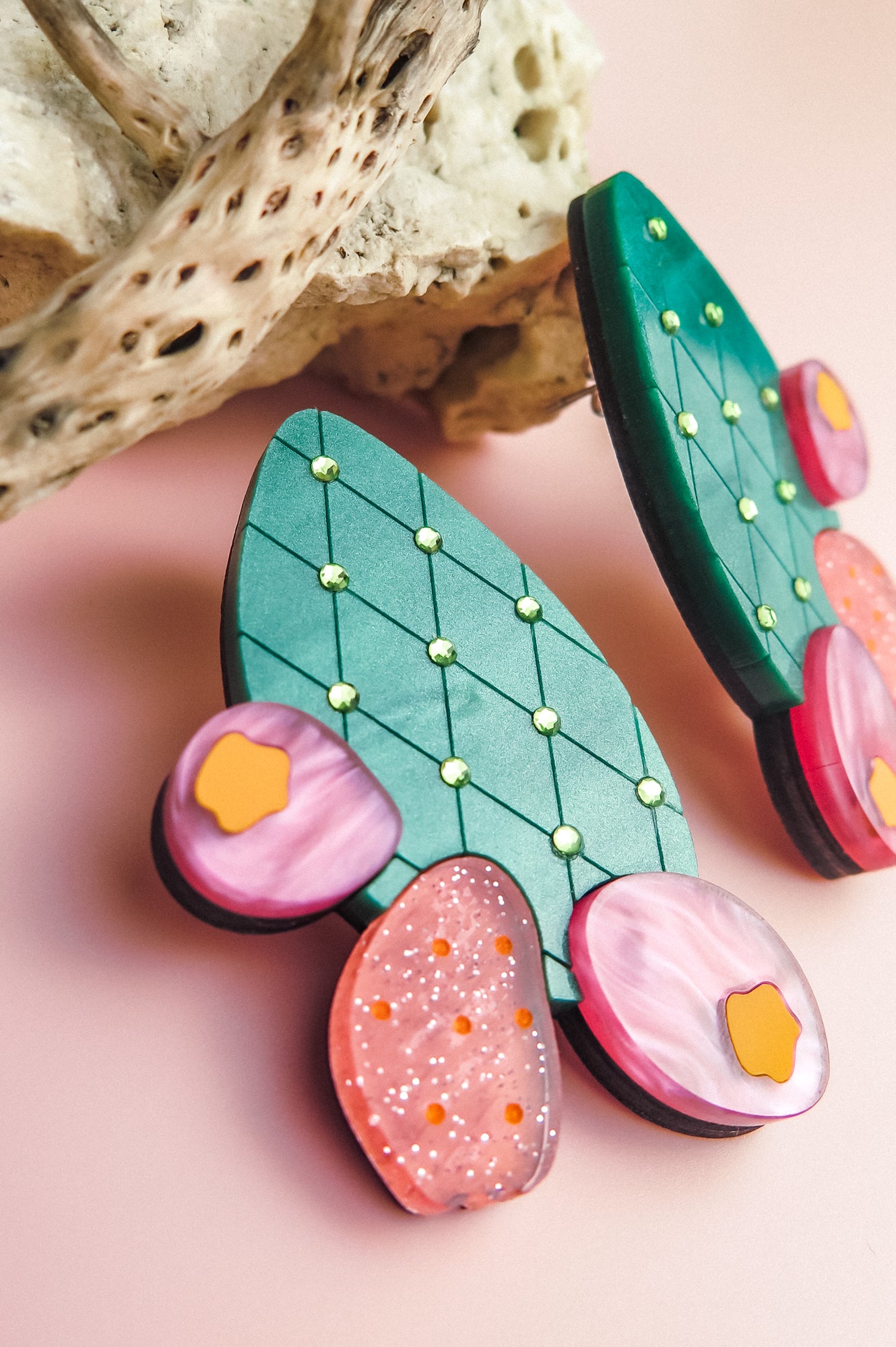 A close-up of colorful, handmade statement earrings shaped like opuncia cactus pads with green, pink, and orange sections, lying on a pink surface next to a textured piece of driftwood.