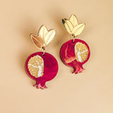 A pair of handcrafted earrings shaped like pomegranates with gold leaves on top and intricate white and gold seed patterns on a red background, displayed on a beige surface, showcasing expert craftsmanship.
