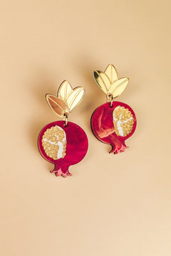 A pair of handcrafted earrings shaped like pomegranates with gold leaves on top and intricate white and gold seed patterns on a red background, displayed on a beige surface, showcasing expert craftsmanship.