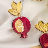 A decorative, handcrafted earring shaped like a pomegranate with golden seeds and a gold leaf at the top, displayed on white fabric with dried grass nearby, showcases expert craftsmanship and attention to detail.
