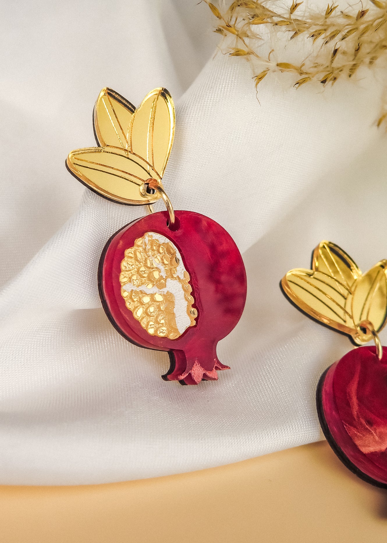 A decorative, handcrafted earring shaped like a pomegranate with golden seeds and a gold leaf at the top, displayed on white fabric with dried grass nearby, showcases expert craftsmanship and attention to detail.