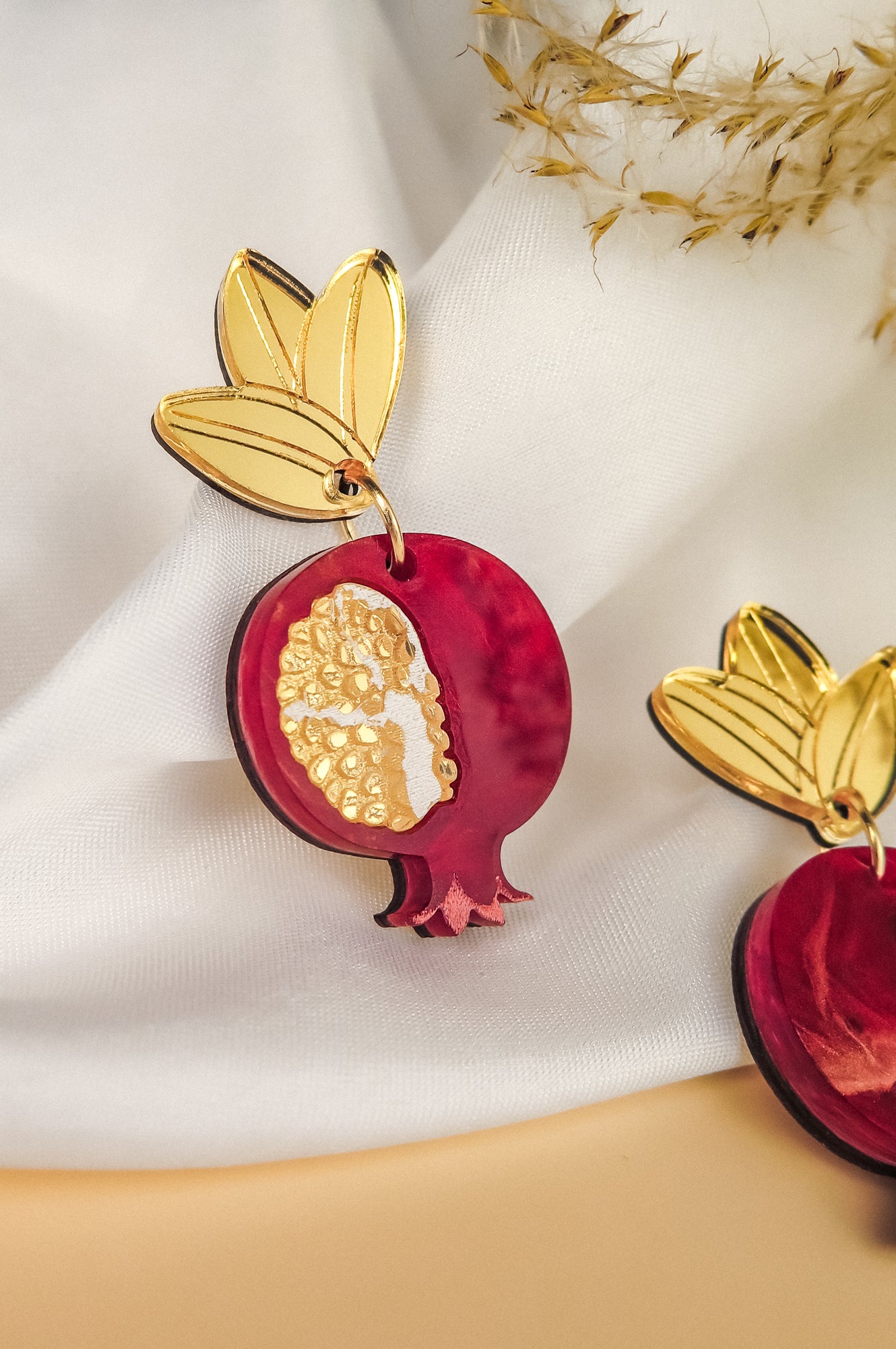 A decorative, handcrafted earring shaped like a pomegranate with golden seeds and a gold leaf at the top, displayed on white fabric with dried grass nearby, showcases expert craftsmanship and attention to detail.
